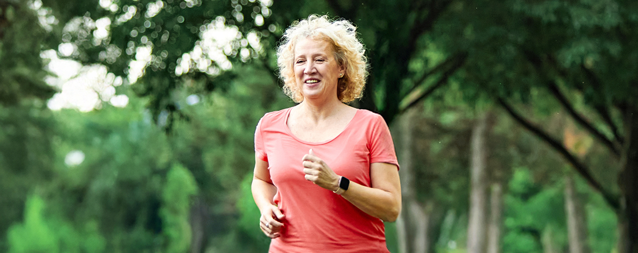 Woman jogging in a park: a woman with curly blonde hair wearing a coral short-sleeved top and a black wristband, running outdoors with trees in the background.